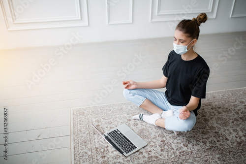 Girl with mask on her face takes yoga classes online sitting on floor with laptop in Lotus position during the quarantine