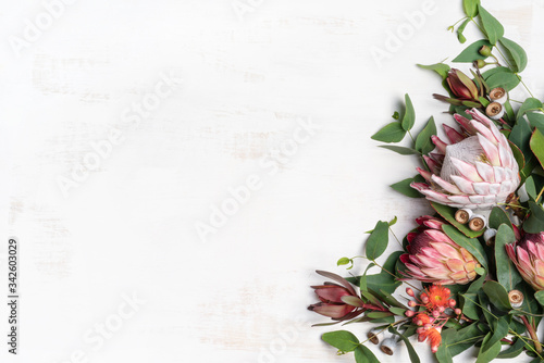 Beautiful pink king protea surrounded by pink ice proteas, leucadendrons, eucalyptus leaves and flowering gum nuts, creating a floral border on a rustic white background.