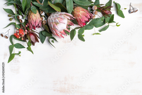 Beautiful pink king protea surrounded by pink ice proteas, leucadendrons, eucalyptus leaves and flowering gum nuts, creating a floral border on a rustic white background.