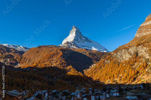 Stunning aerial panorama view of Zermatt town with the famous Matterhorn peak of Swiss Alps in background and golden forest on sunny autumn day with blue sky, Valais, Switzerland