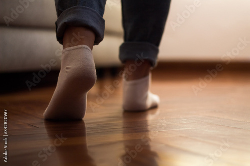 Legs of a woman in jeans and socks walking on the wooden floor of her house with a sofa in the background.