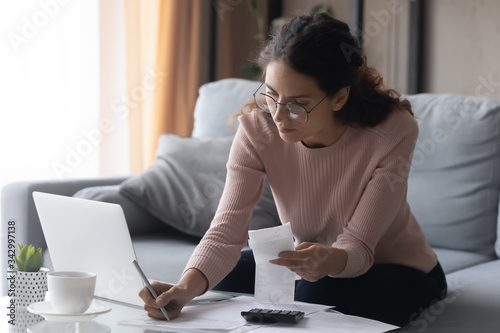 Focused millennial woman in eyewear writes out information from invoices, calculating monthly expenses alone at home. Young lady managing incomes and outcomes, planning investments or summer vacation.