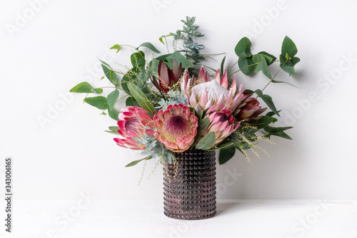 A elegant floral arrangement in a rustic brown vase on a table with a white background. Flower bunch includes a pink king protea, pink ice proteas, leucadendrons, wattle leaves and eucalyptus leaves.