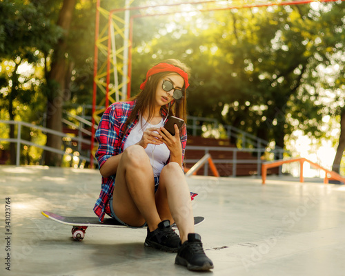 Young stylish woman dressed in youth clothes sits on a skateboard and listens to music on headphones at a skatepark. Summertime