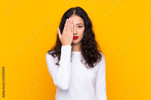 Mixed race woman over isolated yellow background covering a eye by hand