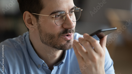 Close up head shot satisfied man wearing glasses holding smartphone near mouth, recording voice message, confident young male chatting online or speakerphone, activating digital assistant