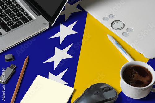 Bosnia Herzegovina national flag on top view work space of creative designer with laptop, computer keyboard, usb drive, graphic tablet, coffee cup, mouse on wooden table.