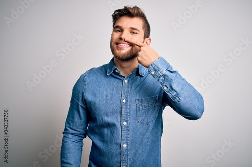 Young handsome blond man with beard and blue eyes wearing casual denim shirt Pointing with hand finger to face and nose, smiling cheerful. Beauty concept
