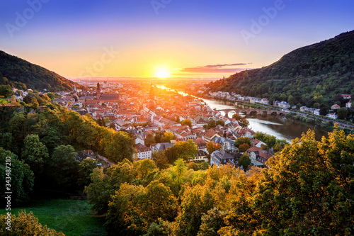 Travel shot: aerial view of Heidelberg, Germany, in beautiful sunset light, framed by colorful trees and gold and purple clear sky, with the Neckar river leading to the sun