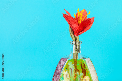 handmade artificial paper red flower in handpainted glass brandy bottle on aquamarine pastel color background