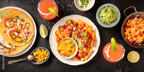 Mexican food, a flat lay panorama on a dark background. Nachos, tortillas, Paloma cocktails, guacamole and other dishes, shot from above