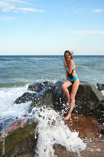 A young girl sits on stones at the sea, splashing waves, waves are breaking on stones, emotions of delight and happiness.
