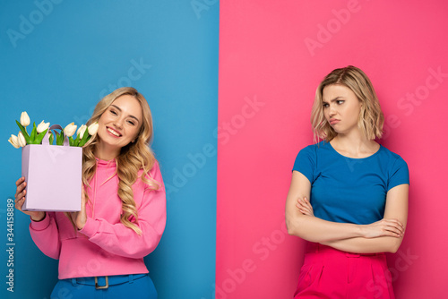 Smiling girl holding bouquet near envy blonde sister on blue and pink background