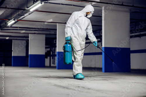 Worker in sterile uniform and mask walking trough underground garage and sterilizing surface. Protection from corona virus / covid-19 concept.