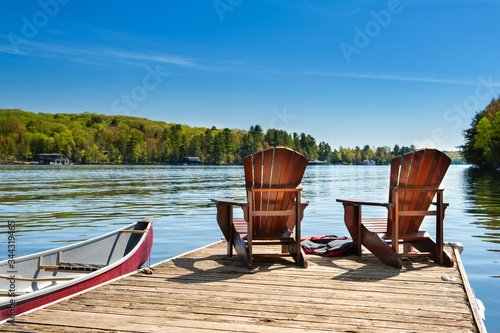 Two Adirondack chairs on a wooden dock on a lake in Muskoka, Ontario Canada. A red canoe is tied to the pier. Across the water cottages nestled between green trees are visible.