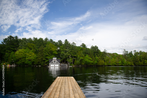 Wooden dock on a calm lake in Ontario, Canada. A Boat house, nestled between green trees, is visible across the water. A plane is crossing the blue sky.