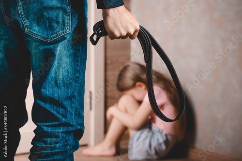 A man with a belt in his hand threatens a little girl. A little girl sits in a corner and hides her face . The concept of domestic violence against a child.