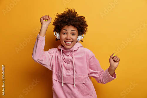 Happy girl has fun, enjoys listening music, relaxes with favorite track, wears wireless headphones, dances with hands raised, wears velvet hoodie isolated over yellow background. Great song lifts mood