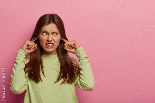 Resentful annoyed woman plugs ears, irritated with loud noise, avoids annoying sound, dressed in casual green jumper, clenches teeth, frowns face, isolated on pink background, blank space aside