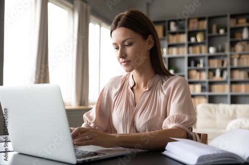 Focused woman journalist freelancer working online on laptop, sitting at desk at home, looking at screen, typing, serious young female writing blog or chatting with friends in social network