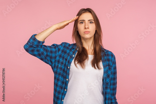 Yes sir! Portrait of patriotic confident young woman in checkered shirt saluting to commander with attentive responsible look, ready to obey order. indoor studio shot isolated on pink background