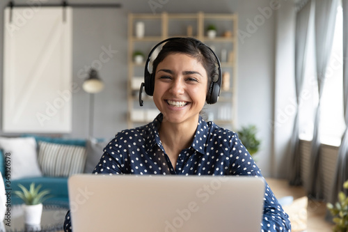 Head shot cheerful smiling pretty hindu ethnic girl sitting at table with computer, wearing headphones with mic, looking at camera. Happy indian woman professional tutor educating client online.