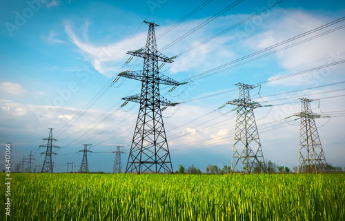 High voltage lines and power pylons in a flat and green agricultural landscape on a sunny day with clouds in the blue sky. Cloudy and rainy. Wheat is growing