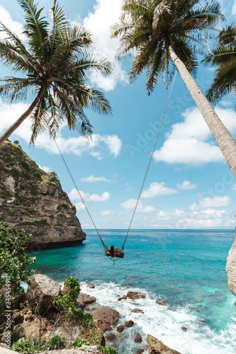 Young girl swinging on a swing overlooking the blue sea. Travel adventure on paradise tropical island Nusa Penida. A young girl swinging on a swing between palm trees on the beach of a tropical island