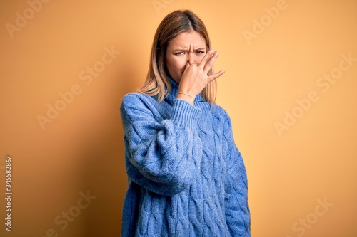 Young beautiful blonde woman wearing turtleneck sweater over yellow isolated background smelling something stinky and disgusting, intolerable smell, holding breath with fingers on nose. Bad smell