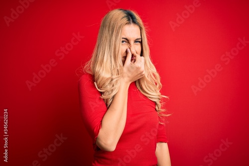 Young beautiful blonde woman wearing casual t-shirt standing over isolated red background smelling something stinky and disgusting, intolerable smell, holding breath with fingers on nose. Bad smell