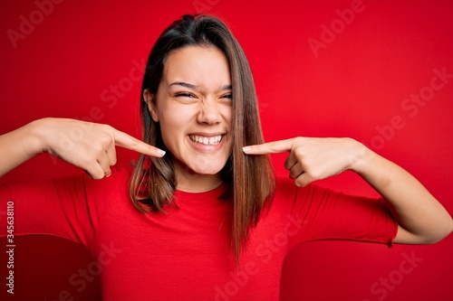 Young beautiful brunette girl wearing casual t-shirt over isolated red background smiling cheerful showing and pointing with fingers teeth and mouth. Dental health concept.