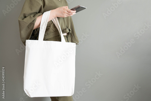 Blank white tote bag canvas fabric with handle mock up design. Close up of woman holding eco or reusable shopping bag and using smartphone near grey wall. No plastic bag and ecology concept.