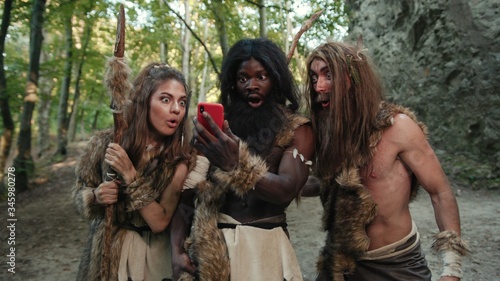 Cheerful group of neanderthal hunters using modern technology smartphone cheering up walking outside cave in the jungle. Technology and first people.