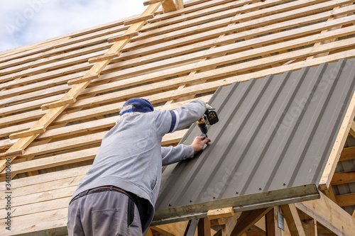 Senior gray-haired Construction man using a screwdriver, fastens a roofing sheet to wooden rafters on the roof of a country house under construction. Physical activity of the seniors.