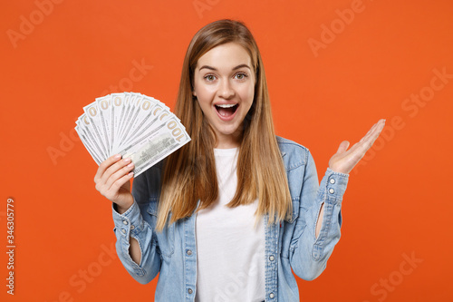 Surprised young woman girl in casual denim clothes posing isolated on orange background. People lifestyle concept. Mock up copy space. Holding fan of cash money in dollar banknotes, spreading hands.