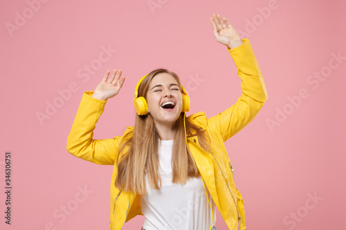 Funny young woman girl in yellow leather jacket posing isolated on pastel pink background in studio. People lifestyle concept. Mock up copy space. Listen music with headphones, rising hands, dancing.