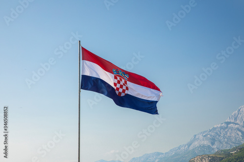 The red white blue Croatian flag waving in the wind on a flagpole on a sunny day and a blue sky, with in the background the Biokovo mountains on Makarska, Dalmatia at the Mediterranean coast