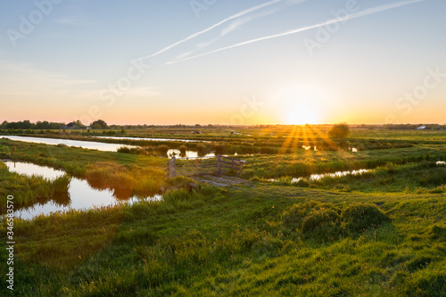 Sun sets over the wide open Dutch polder landscape. Green meadows intersected with ditches filled with water.