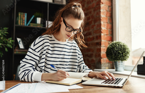 Happy millennial woman taking notes in notepad while working at laptop in comfortable loft office.