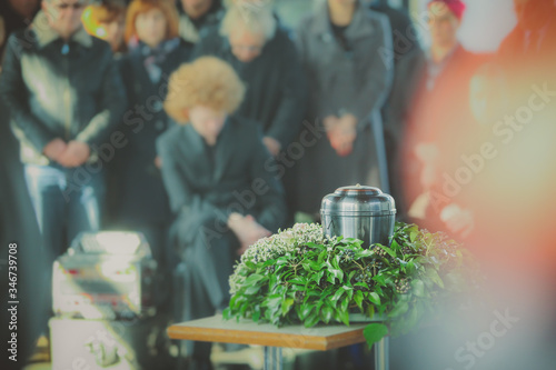 A metal urn with ashes of a dead person on a funeral, with people mourning in the background on a memorial service. Sad grieving moment at the end of a life. Last farewell to a person in an urn.