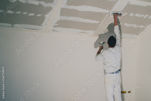 Man dressed in white, a painter, is applying putty or stucco to a dry wall installation in a house. Smoothing surfaces of a drywall application, reaching high up to the roof.