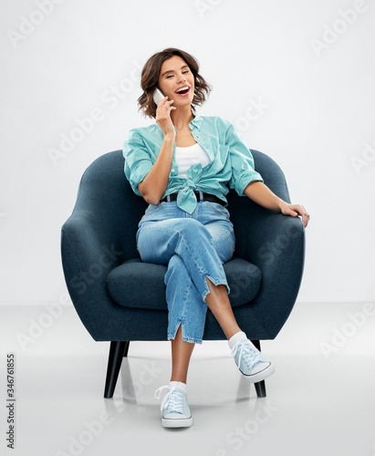people and technology concept - portrait of smiling young woman in turquoise shirt and jeans sitting in modern armchair and calling on smartphone over grey background