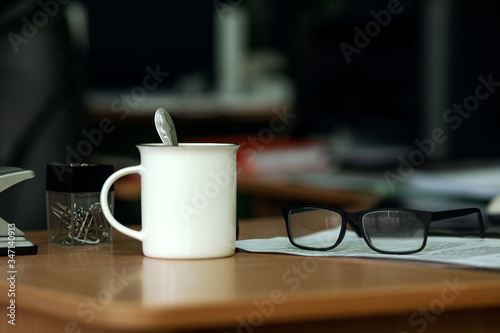a white coffee mug, black-rimmed glasses, a white sheet of printed text, and a clear container of paper clips on a wooden office Desk