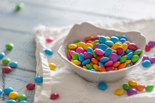 Smarties put in a white porcelain bowl and scattered around on a white clothe and a table