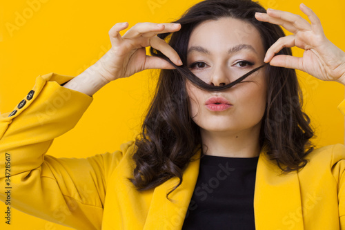 Stylish young woman in a yellow jacket on a yellow background, making a mustache out of her hair.