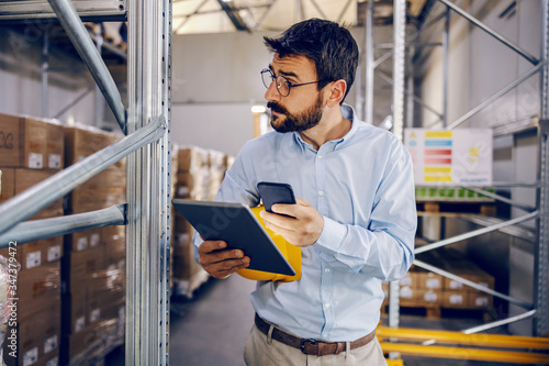 Young handsome bearded employee holding tablet and smart phone in hands and trying to finish work on time. Warehouse interior.