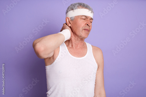 Studio shot of mature man with painful neck, male posing isolated over lilac background with hand on his neck, posing with closed eyes, wearing white t shirt and head band. Health care, sport injury.