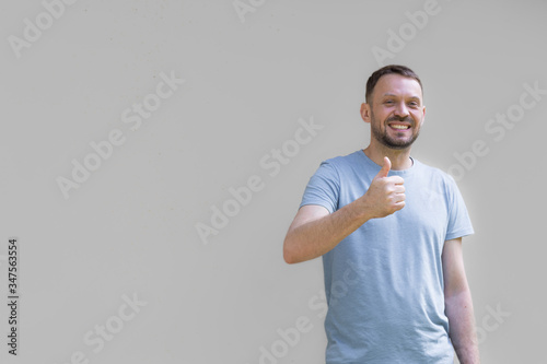 Uśmiechnięty mężczyzna na szarym tle, pokazujący dłonią gest ok, uniesiony w górę kciuk