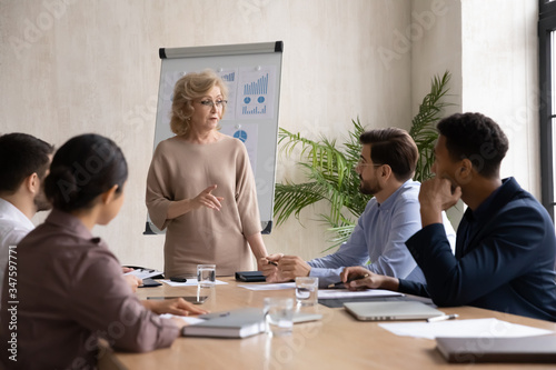 Confident mature 60 year old businesswoman presentation new project in boardroom at company meeting. Adult woman coach auditor speaks about business on flip chart graphs background.