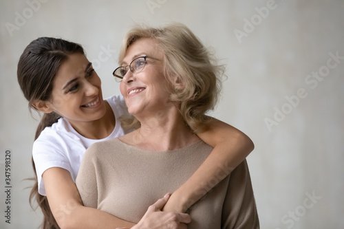 Close up headshot portrait picture of adult daughter hugs from behind happy mature mother enjoying tender moment looking at each other. Smiling woman and older mum embracing having fun together.